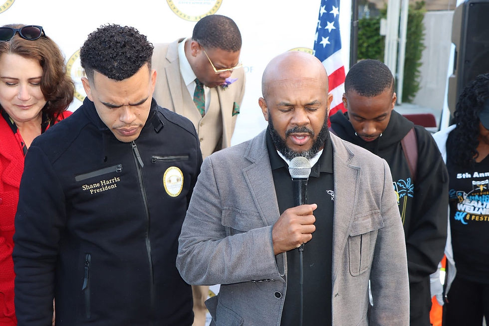 Pastor Glenn McKinney leads a prayer as one of Shane Harris’s Gas Me Up community events gets underway. Standing beside him, community members bow their heads in reflection and unity. Shane Harris, wearing a black jacket embroidered with “President,” stands to McKinney’s left. The moment captures a spirit of faith, service, and togetherness at the event. March 2022.