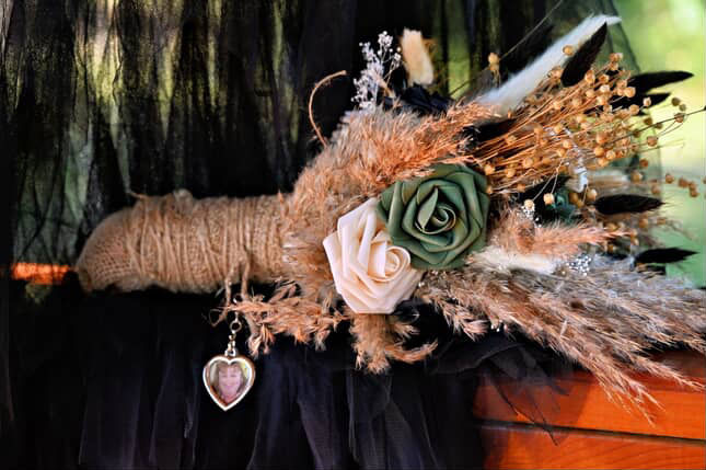 Bouquet with green and beige roses, dried plants, and a heart locket against black tulle and wood. Nature-themed, rustic vibe.