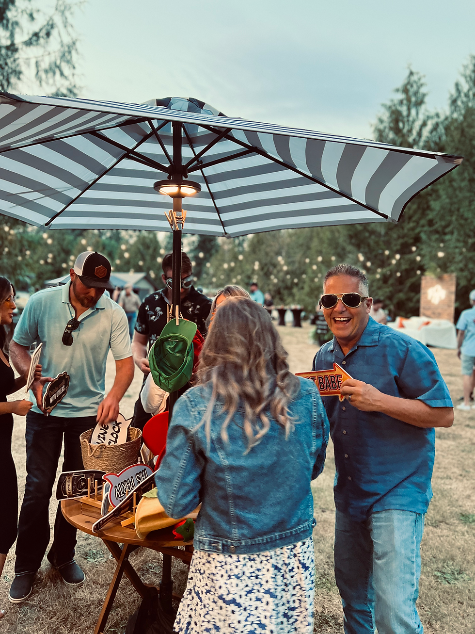 Adults gathering around an outdoor table filled with funny photo booth props, laughing and having fun