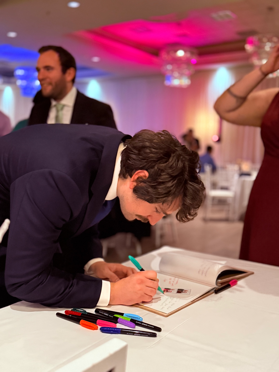 A gentleman wearing a suit leaning down to sign a guest book full of photo booth strips at a table in a room with colored lights and people mingling and having fun