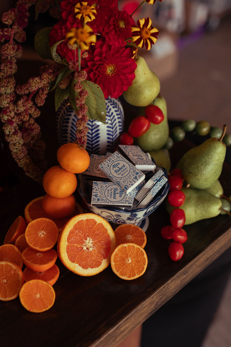 Vibrant arrangement with red flowers, oranges, pears, and tomatoes. Blue patterned vase holds "Alex Robert" matchboxes. Warm and lively.