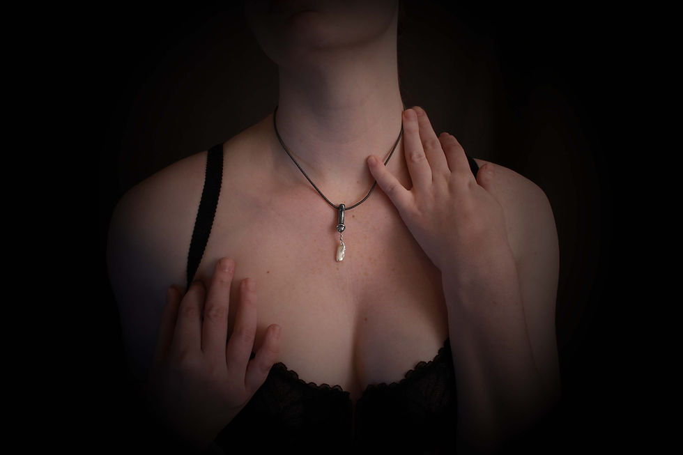 Thumbnail: Penis pendant with large pearl on black cord on woman's neck. She wears a black bra and one hand is on her shoulder.