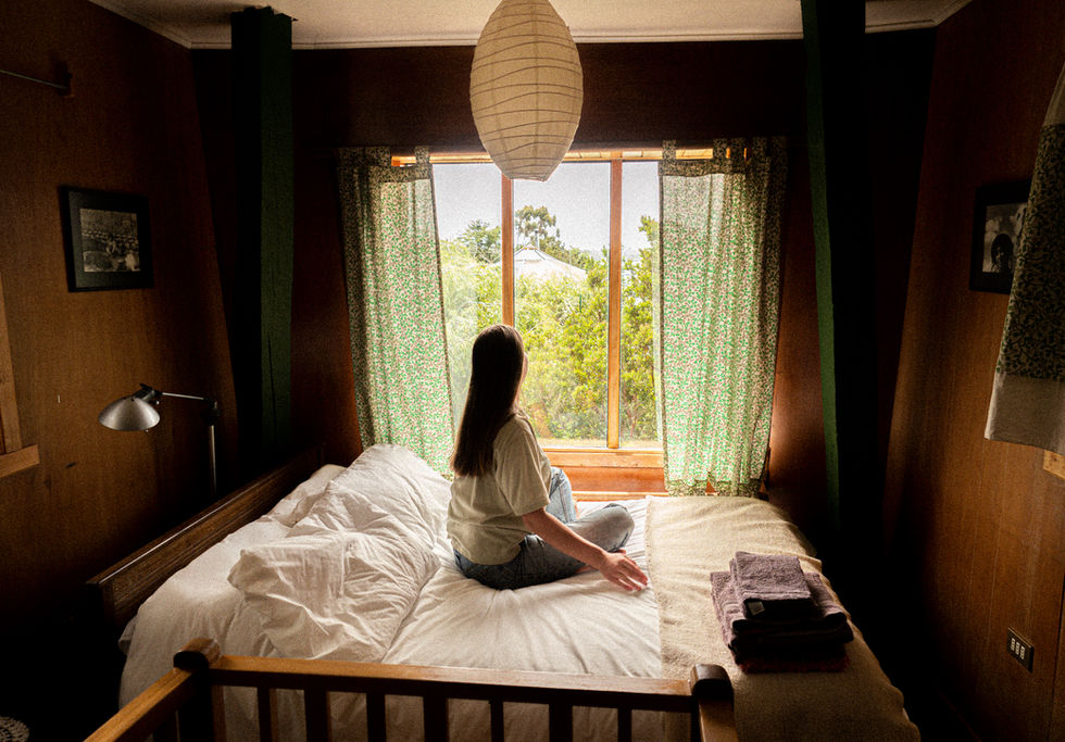 Oceanside lodging on Chiloe- woman sitting on a bed looking out a window at trees and the ocean