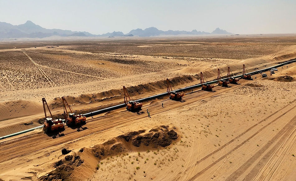 A high-angle, aerial shot of a long industrial pipeline stretching across a vast, sandy desert landscape. Several dark shapes, likely service vehicles or equipment, are positioned alongside the pipe at regular intervals under a bright sky.