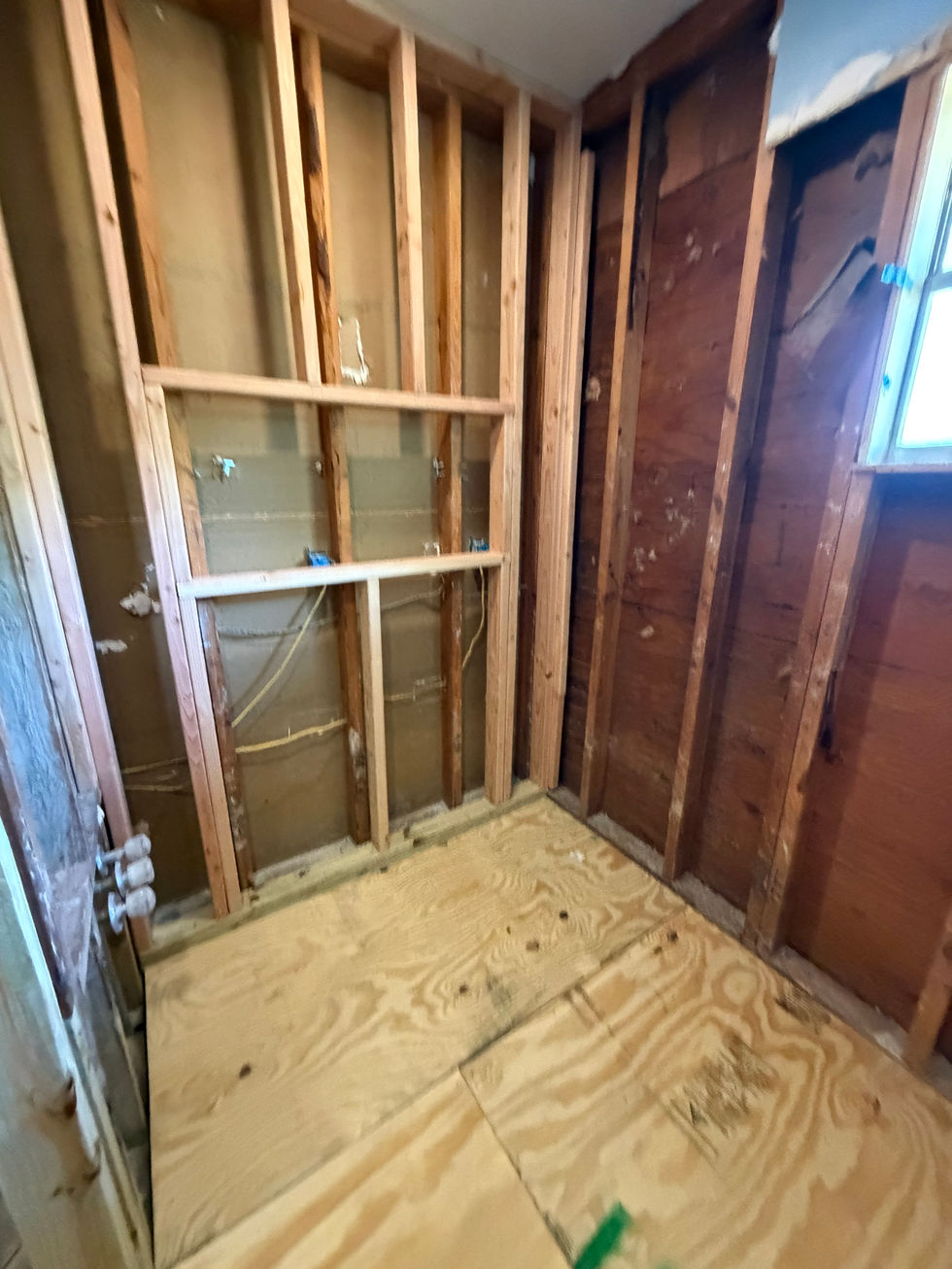 Bathroom corner with exposed studs, horizontal blocking, and fresh plywood flooring.