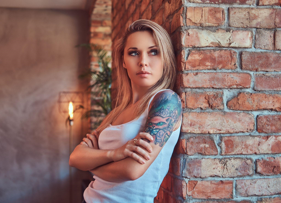 A woman with colored tattoo inks on her arm leaning on a brick wall
