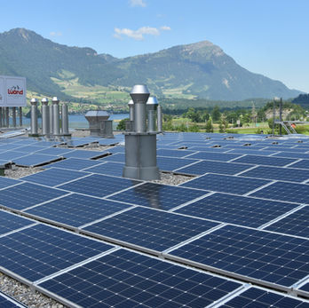 Zahlreiche Solarmodule auf einem Dach vor Berg- und Seenlandschaft.