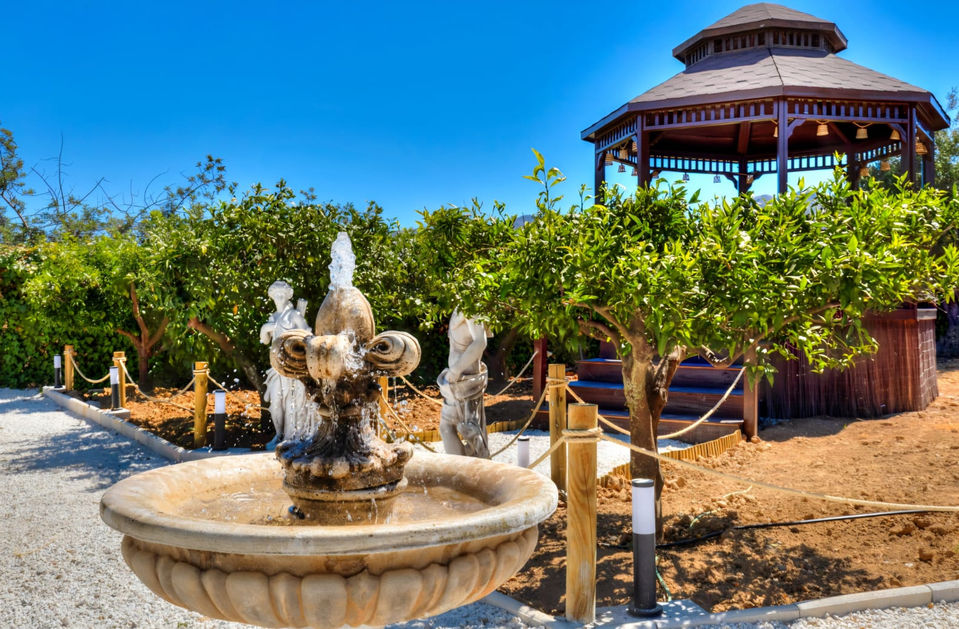 an outdoor garden scene featuring a stone water fountain with sculptures and water jets in the foreground. Behind the fountain, there are green leafy trees, a wooden gazebo with a hexagonal roof, and a gravel pathway bordered by rope fencing.