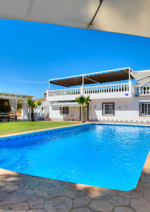 a bright outdoor pool area at a white two-story house with a covered balcony and large umbrellas providing shade. The scene includes cushioned lounge chairs around the pool, a pergola with draped curtains on a grassy lawn, and clear blue sky indicating a sunny day.