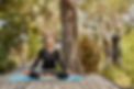 Cindy Robins is sitting on a yoga mat on a wooden deck in an outdoor area with trees and bushes around.