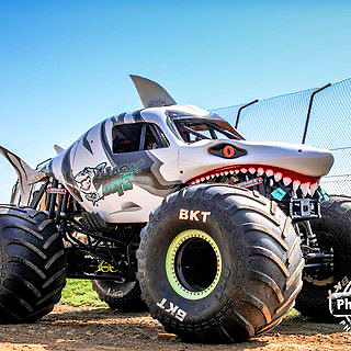 A large, shark-themed monster truck is parked in a dirt lot.