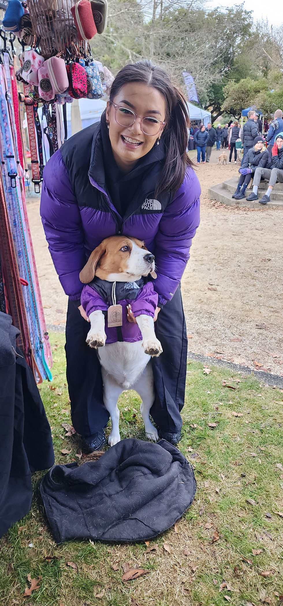 A customer at a Hendricks and Maple market stall holding up her Beagle, both wearing matching purple and black puffer jackets — the customer in her own North Face jacket, the Beagle in a matching Hendricks and Maple 'The Dog Face' puffer jacket with brand tag.
