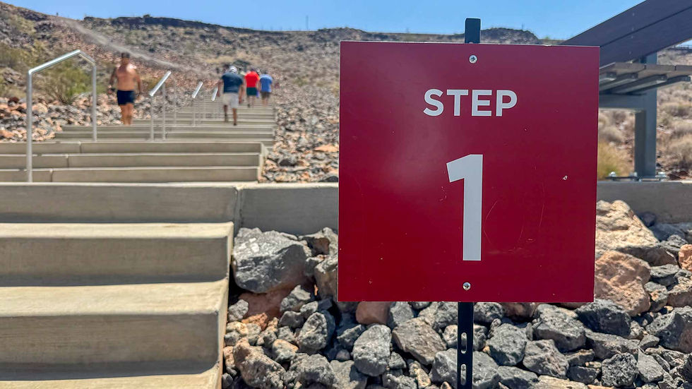 People walking up rocky outdoor stairs under blue sky. Foreground shows red sign with "Step 1" text. Rugged landscape in background.