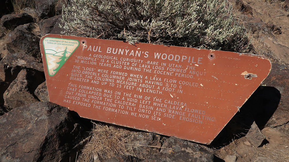 Sign titled "Paul Bunyan's Woodpile" explains geological formation of lava logs, set against rocky terrain with dry vegetation.