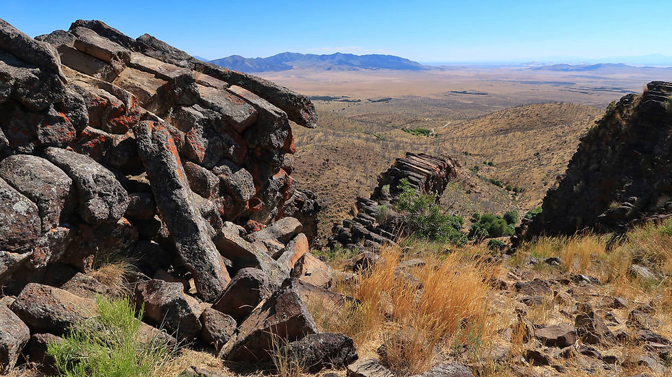Rocky ridge with textured boulders in the foreground, overlooking vast dry plains and distant mountains under a clear blue sky.
