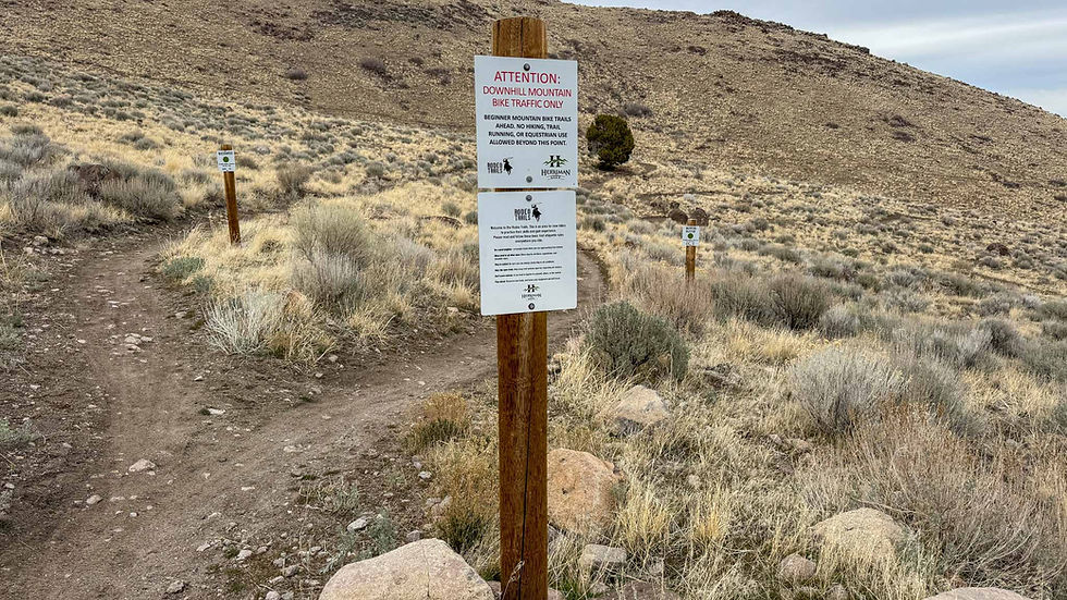 Trail sign warns of "Downhill Mountain Bike Traffic Only" in a dry, grassy landscape with a dirt path and distant hills.