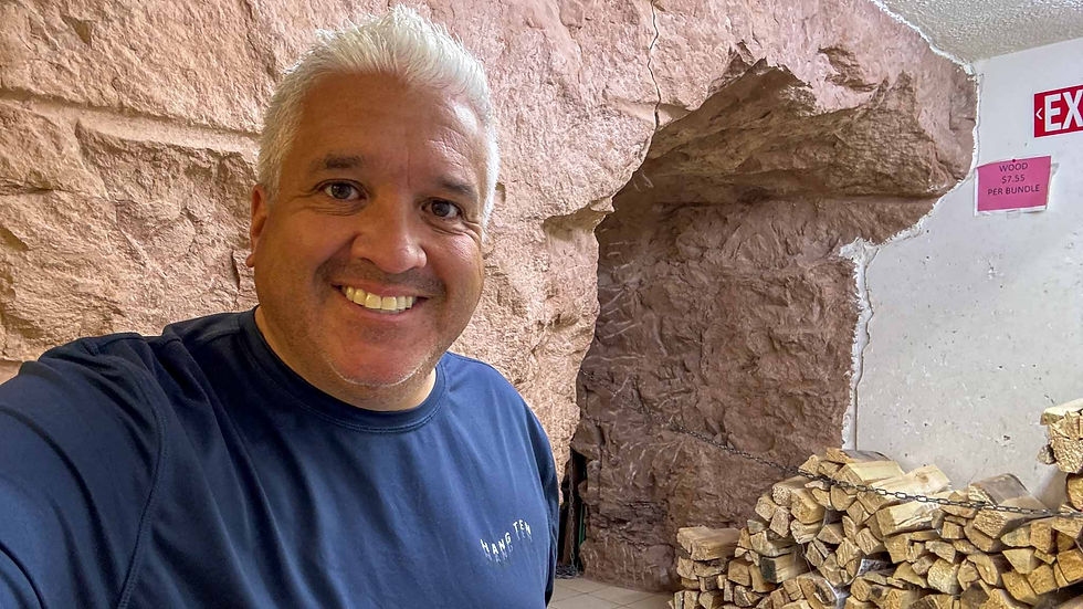 Smiling man in navy shirt selfie inside a rock-walled room beside stacked firewood, with an EXIT sign and wood price sign.