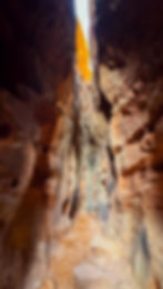 Narrow rocky canyon with tall, textured red and brown walls. Sunlight peeks through the top, casting a warm glow on the path below.