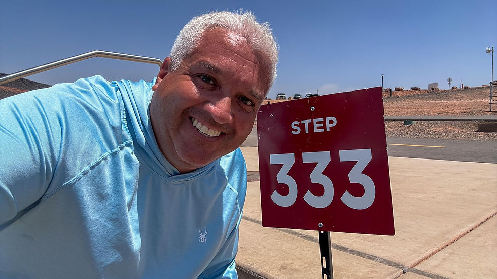 Smiling man in blue shirt next to a red sign reading "Step 333" on a sunny day. Desert-like landscape in the background.