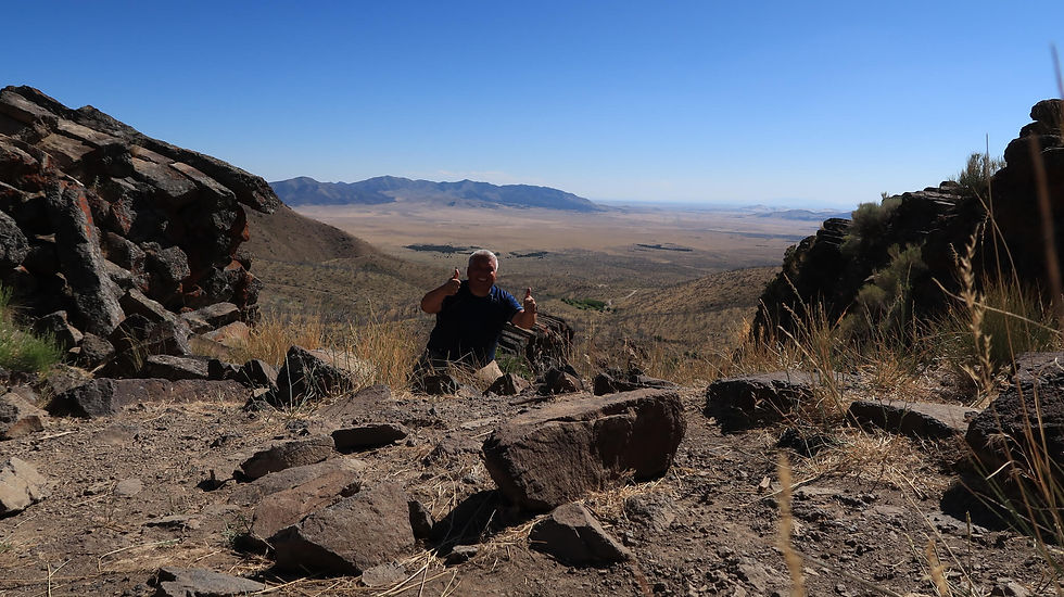 Man giving thumbs up in a rocky desert landscape with mountains in the distance, under a clear blue sky, conveying a joyful mood.