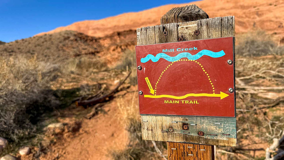 Red Mill Creek trail sign with MAIN TRAIL arrow in a desert landscape of orange hills and blue sky.