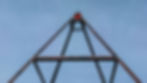 Looking up at a tall metal pyramid structure against a clear blue sky, a person in red appears at the top, evoking adventure and scale.