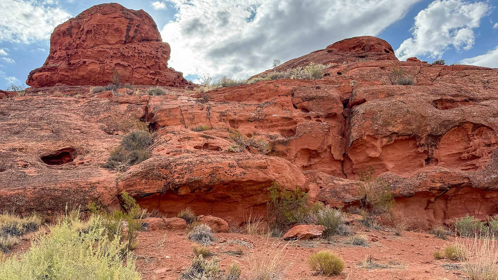 Red rock formations under a partly cloudy sky with scattered desert vegetation in the foreground, creating a rugged and arid landscape.