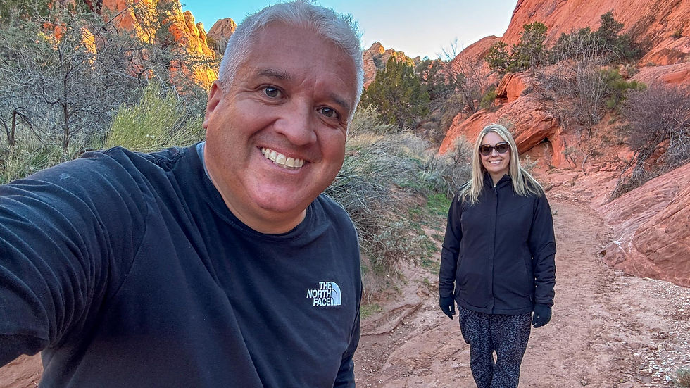 A man and woman hiking on a red rock trail, smiling. The man wears a black North Face shirt. Clear sky and desert landscape in the background.