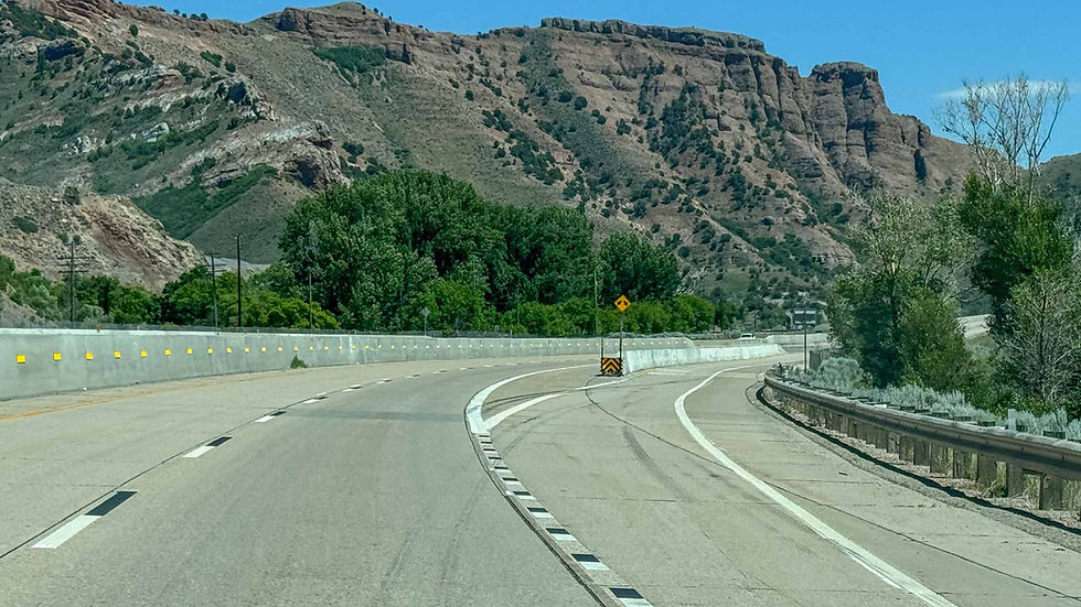 Empty curving highway through rocky desert mountains under a clear blue sky, with green trees and road signs