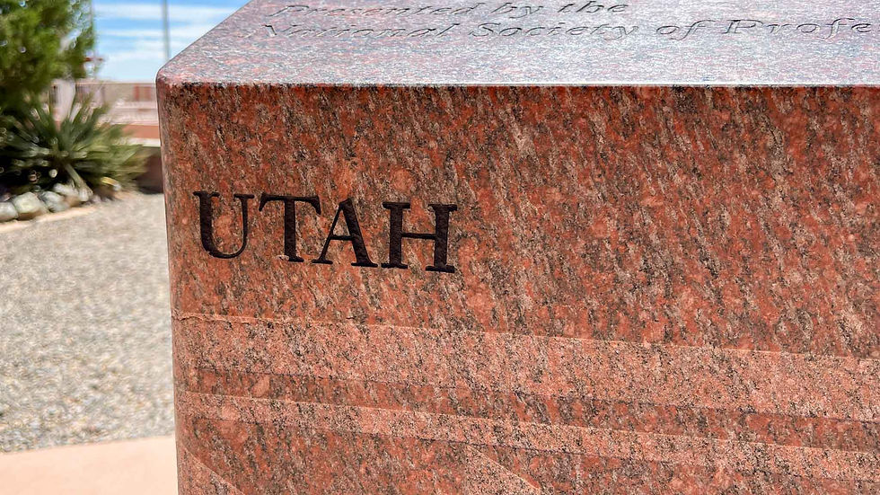 Red granite block with the word "UTAH" engraved. Background shows a garden with gravel, green plants, and a blue sky. Text is partially visible.