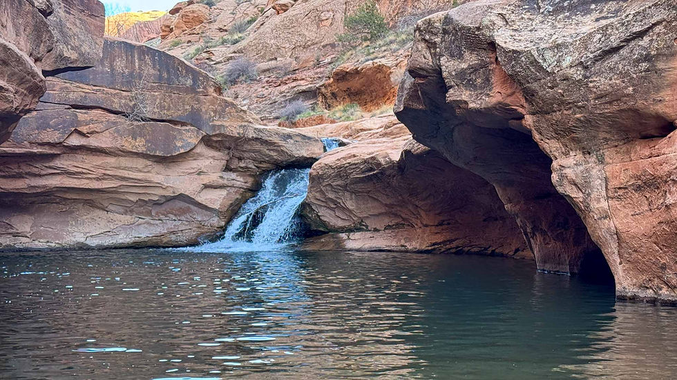 Small waterfall cascades between red canyon rocks into a calm dark pool under a rugged desert overhang.