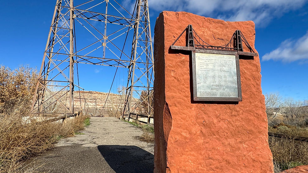Rusty bridge and red stone monument with plaque under a clear blue sky. Dry foliage and distant cliffs in the background.
