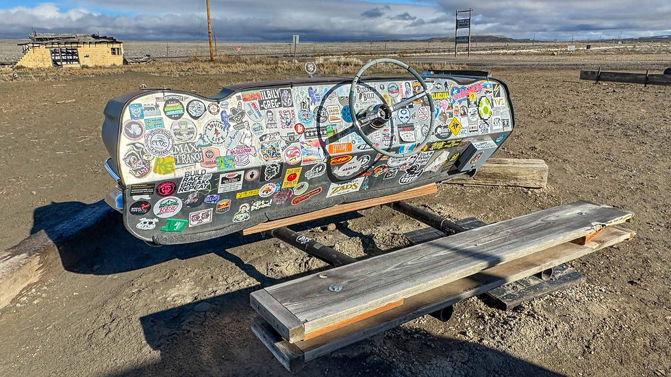 Steering wheel bench covered in stickers, placed outdoors on dirt. Wooden planks in front, abandoned structure in background, clear sky.