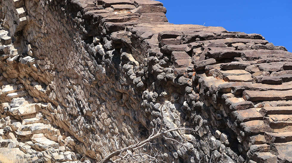 Close-up of a rugged stone wall with dark and light rocks against a clear blue sky. A dry branch is in the foreground.