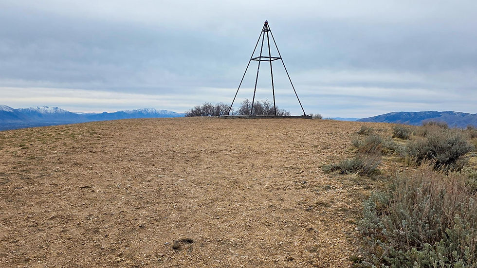 Rocky hill with sparse shrubs and a metal pyramid structure. Snowy mountains under a cloudy sky in the background.