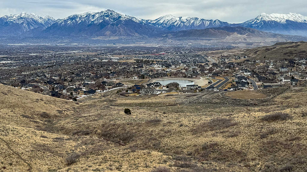 Aerial view of a sprawling town with houses, surrounded by hills. Snow-capped mountains in the background under a cloudy sky.