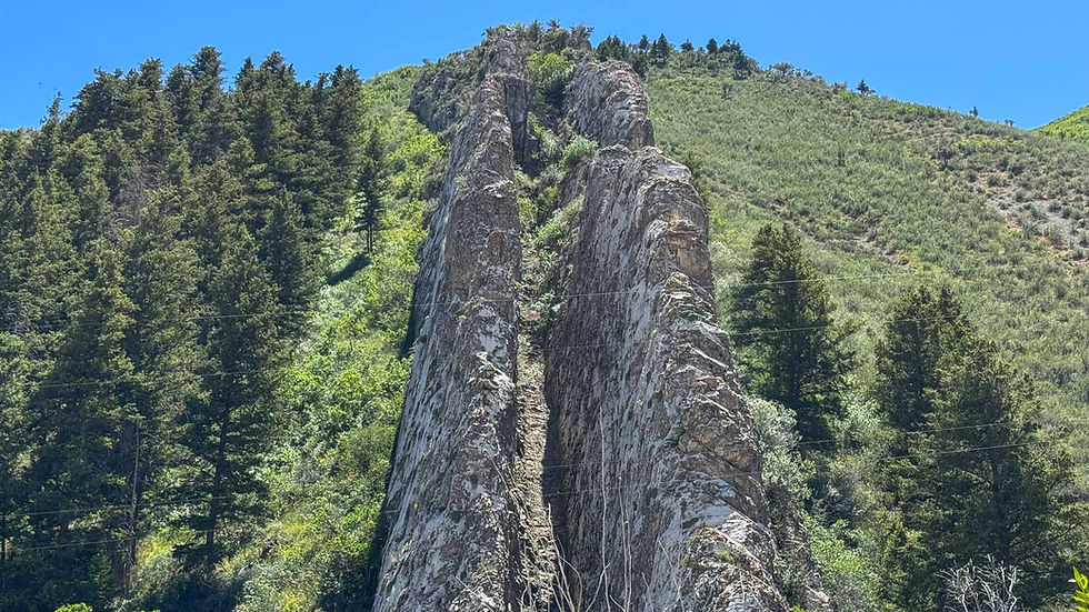 Tall split rock spire rises from a green forested mountainside under a clear blue sky.