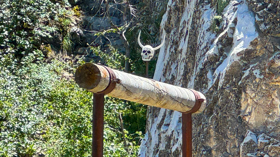 Rusty pipe mounted on posts beside a cliff, with a skull sign in a wooded rocky setting, eerie and rugged.