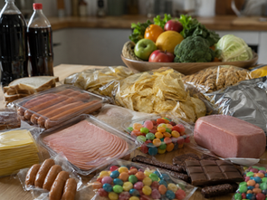 Bord fullt av ultraprocessad mat som korv, skinka, chips och godis i plastförpackningar, med färsk frukt och grönsaker skymtande i bakgrunden.