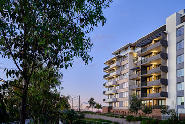 Contemporary mid-rise apartment building at dusk with illuminated balconies and modern façades, positioned along a main road within a masterplanned residential precinct, showcasing premium apartment living, urban design, and high-quality residential architecture.