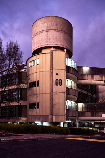 Brutalist concrete building with cylindrical tower and elevated enclosed walkways at dusk, featuring industrial architectural design and curved façade elements. Sydney Architectural photography highlighting modernist civic or institutional structure, exposed concrete forms, and dramatic twilight sky in an urban streetscape setting.