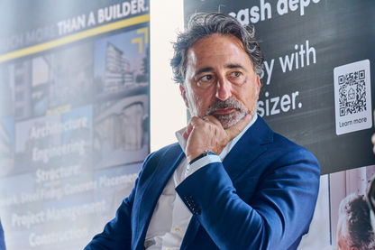 Business executive in a blue suit listening thoughtfully during a corporate event, seated beside promotional signage with a QR code.
