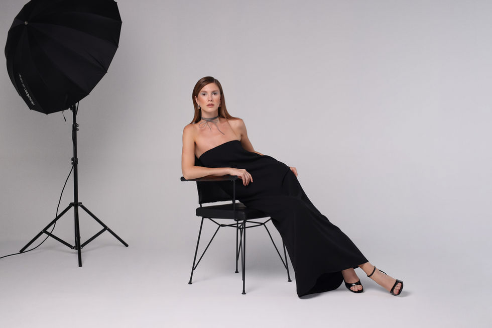 Stüdyo ışık ayağı yanında siyah sandalyede oturan siyah elbiseli kadın model, editoryal moda ve lookbook fotoğrafı.