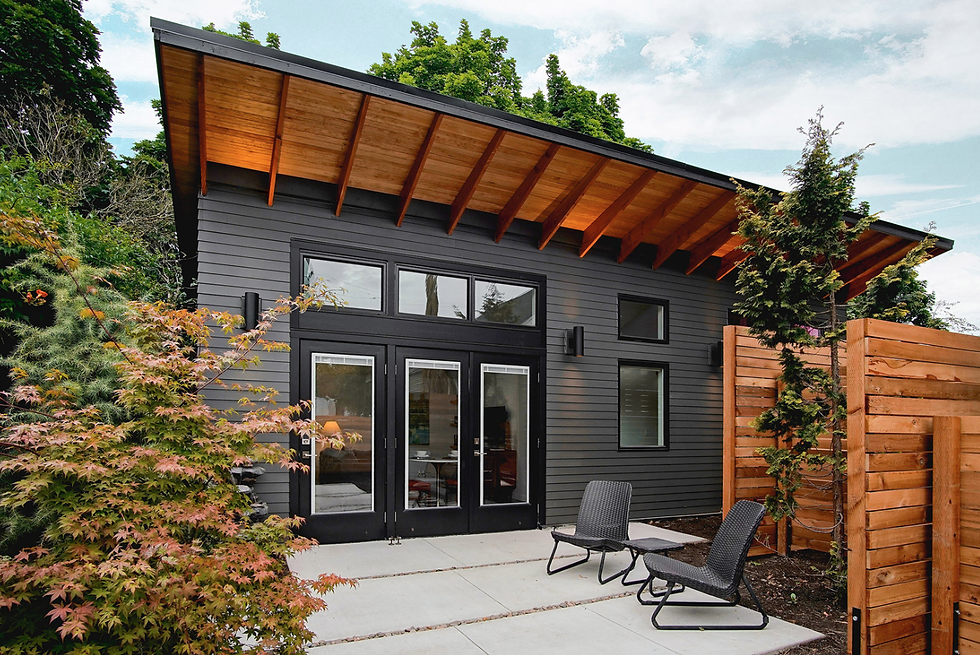 Modern gray house with large glass doors, wooden awning, and patio chairs. Surrounded by trees and a wood fence under a cloudy sky.