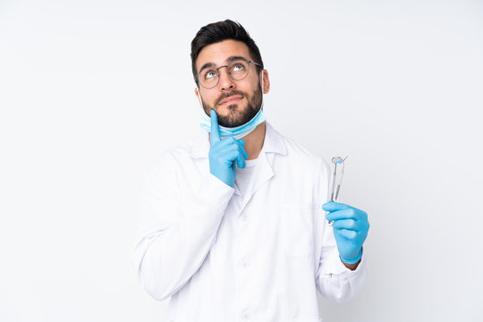 Man in white coat and blue gloves ponders with dental tools in hand, against a plain white background, exuding a thoughtful mood.
