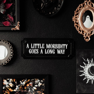 Black and white gothic style wall plaque with text saying "a little morbidity goes a long way"’