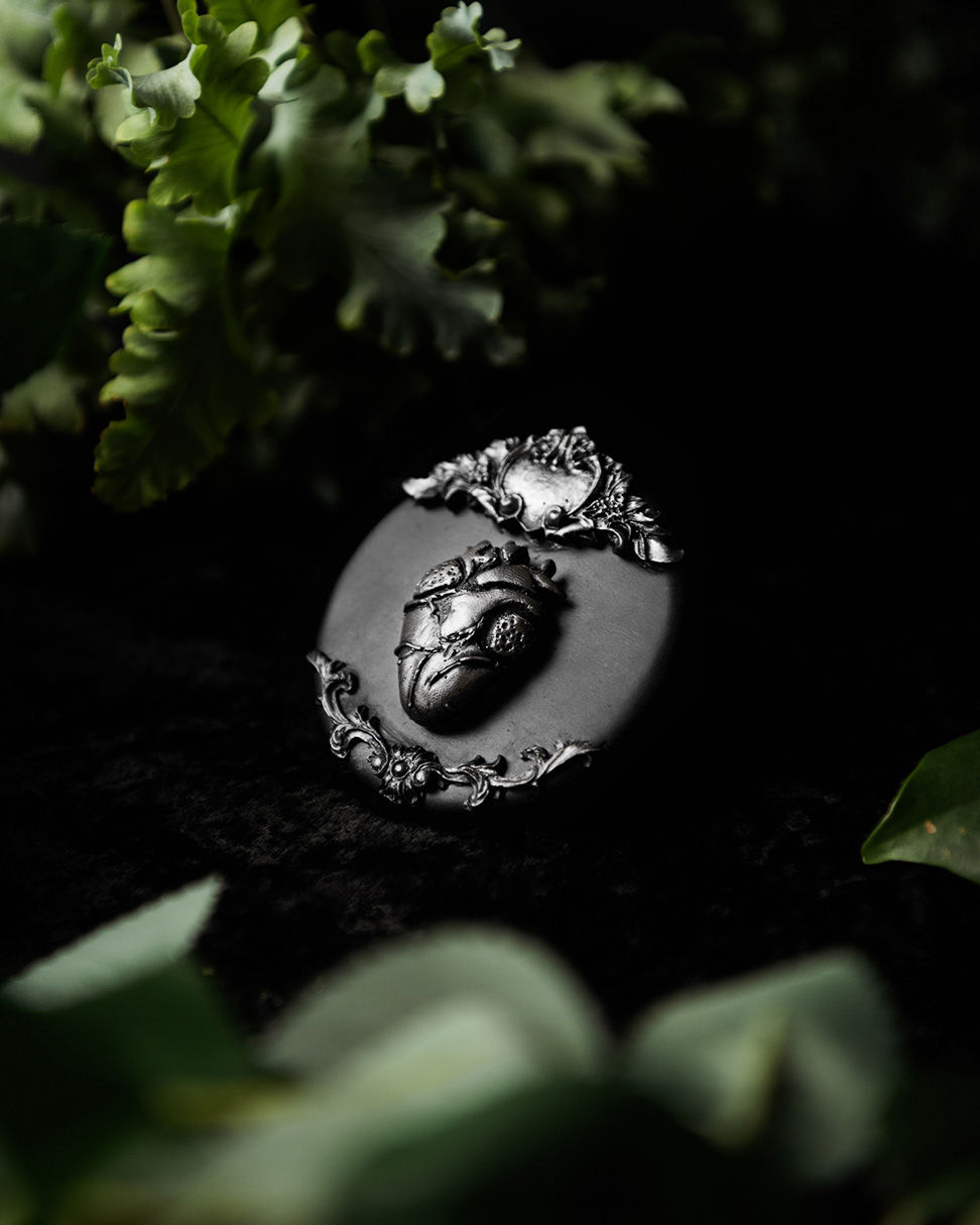 Thumbnail: A round, black wall decor piece with an anatomical heart design in silver and baroque-style flourishes at the top and bottom.