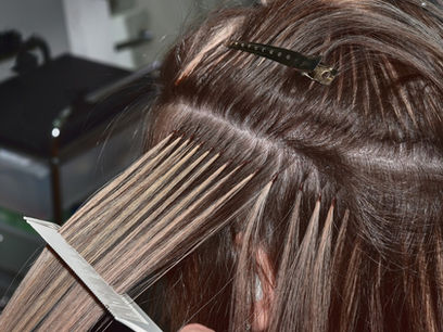 Hair extensions applied to blonde hair using fusion technique. Hands and tools are visible. Hair clips secure sections. Salon setting.