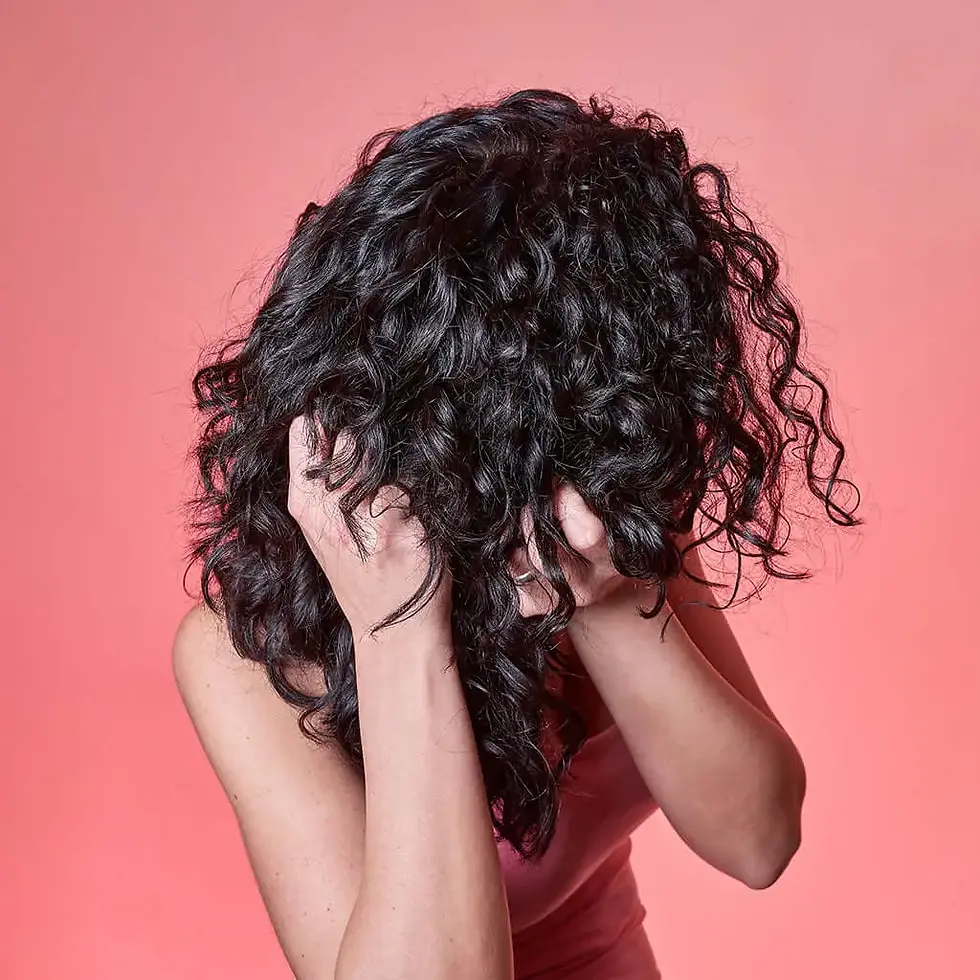 Person with curly black hair covering face with hands against a pink background, conveying frustration or stress.