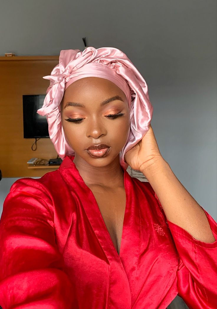 Woman in a red robe with a pink satin headwrap, eyes closed, serene expression. Background shows a wooden shelf and gray wall.